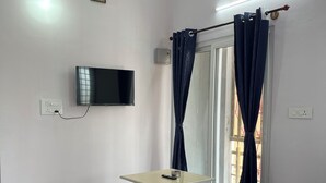 A modern living room with a flat-screen TV, a small table, blue sofas, a glass door with curtains, and a ceiling light fixture.