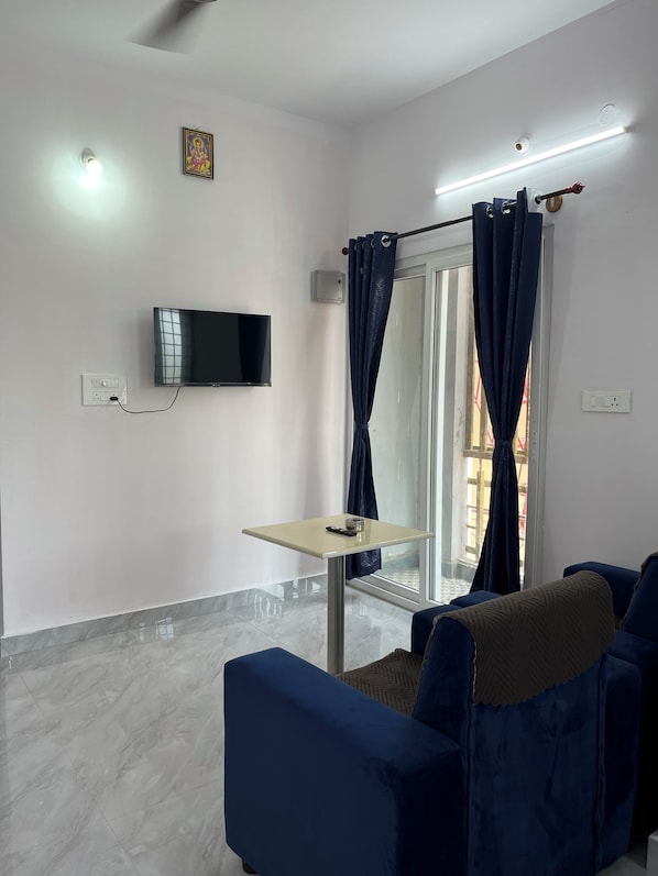 A modern living room with a flat-screen TV, a small table, blue sofas, a glass door with curtains, and a ceiling light fixture.