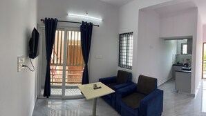 A living area with a sofa, a small table, a TV mounted on the wall, and a balcony with a railing.