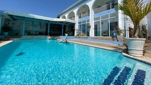 A swimming pool with clear blue water, a covered patio area, and a building with white walls and arches.