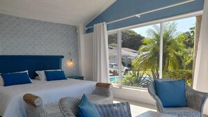 A bedroom with a large bed, two wicker chairs, a small table, and a view of a swimming pool and palm trees.