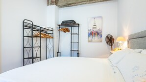 Ein Bett mit weißer Bettdecke, ein Metallschrank mit Kleidung, eine Leiter, ein Ventilator, eine Lampe und ein Bild des Eiffelturms.