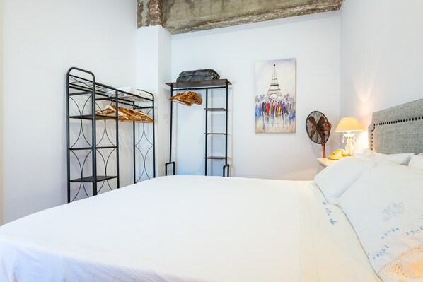 A bed with a white sheet, a metal wardrobe with clothes, a ladder, a fan, a lamp, and a painting of the Eiffel Tower.