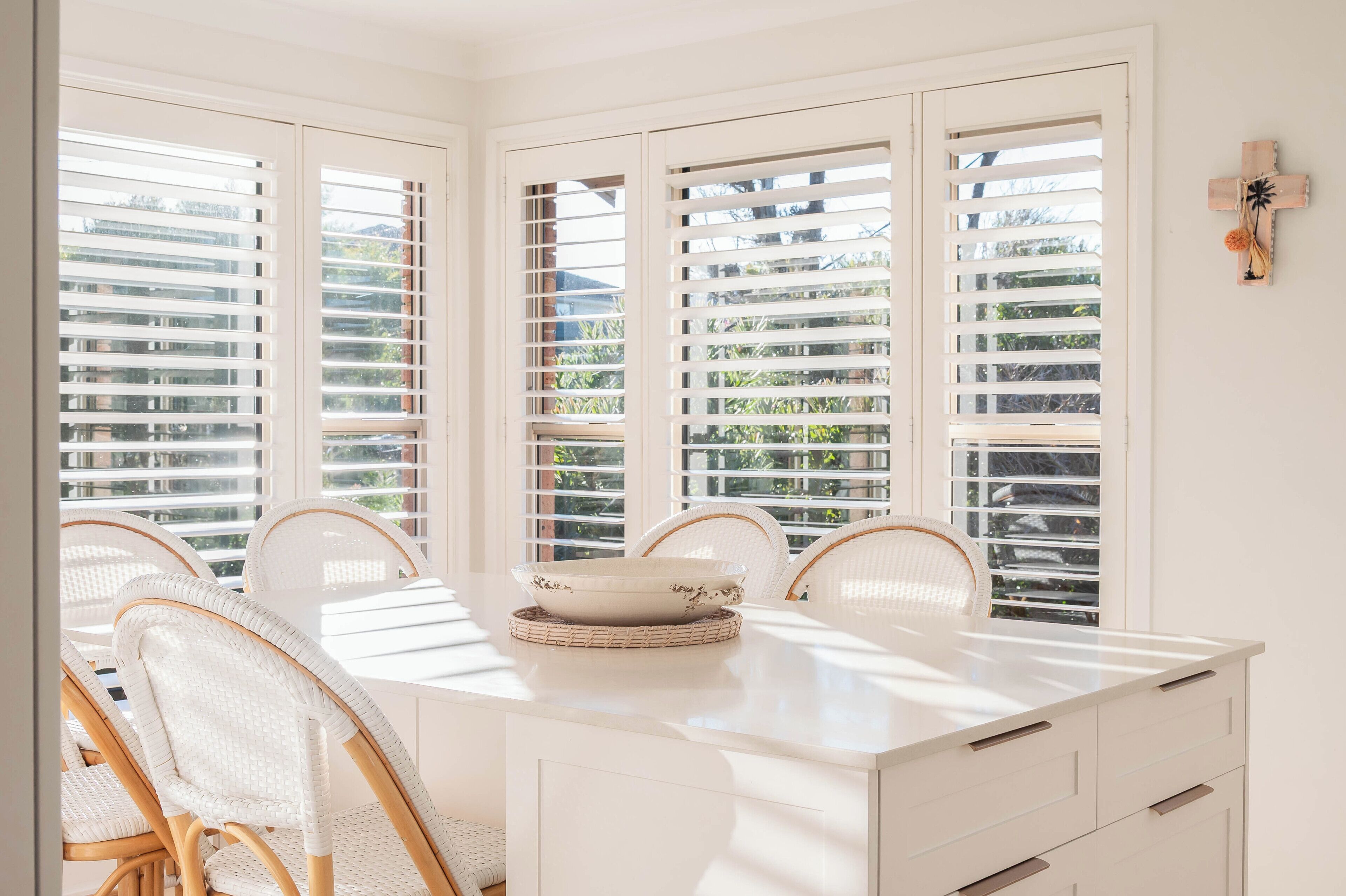 A dining area with a white table, wicker chairs, and a large window with blinds.