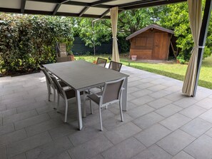 A covered patio with a table and chairs, surrounded by greenery and a wooden structure.