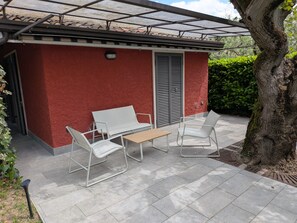 Un patio con un muro rojo, una zona de estar techada y un árbol.