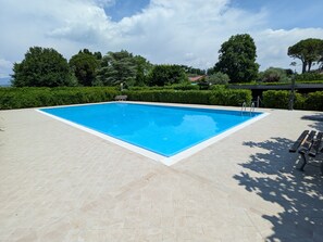 A rectangular swimming pool with a tiled deck, surrounded by greenery and trees.