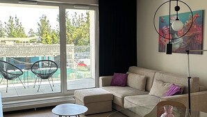 A modern living room with a glass coffee table, a sofa, and a dining table. There is a large window providing a view of the outside.