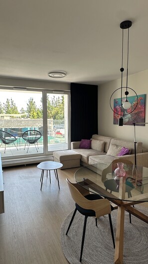 A modern living room with a glass coffee table, a sofa, and a dining table. There is a large window providing a view of the outside.