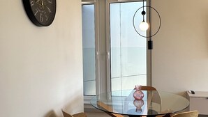 A modern dining area with a glass table, four chairs, a round rug, a wall clock, and a pendant light.
