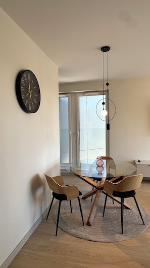 A modern dining area with a glass table, four chairs, a round rug, a wall clock, and a pendant light.
