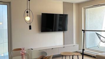 Una sala de estar moderna con un televisor de pantalla plana montado en la pared, una mesa de comedor de cristal rodeada de sillas, un sofá y una lámpara de pie.