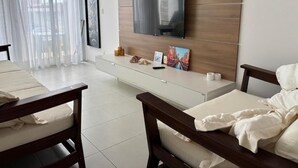 A modern living room with a flat-screen TV mounted on a wooden wall, a white console table with decorative items, a bed with white bedding, and a large window with sheer curtains. - Ipioca Beach House (Maceió)