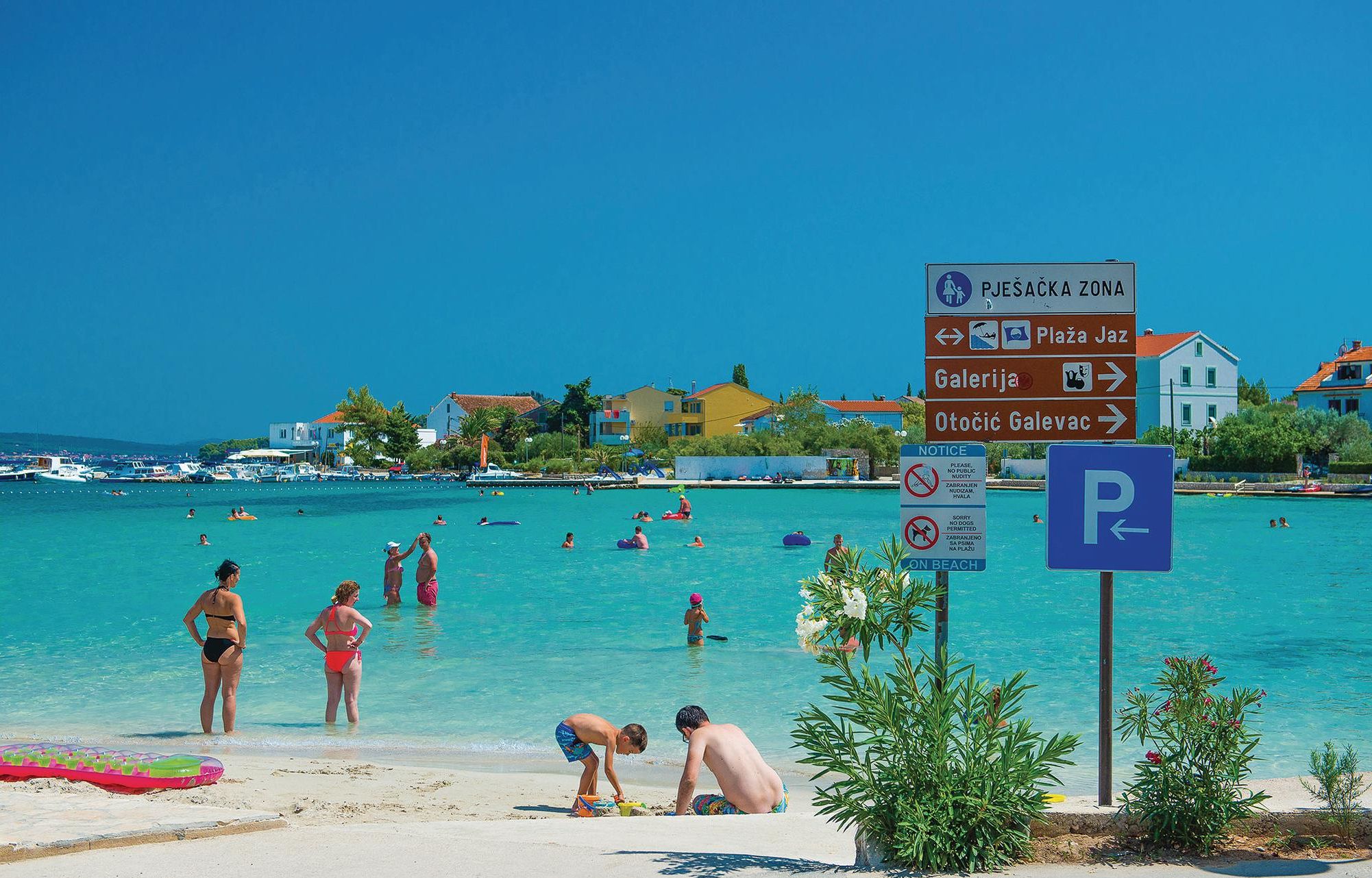 A beach with clear turquoise water, people swimming and sunbathing, and a signpost with directions to local attractions.