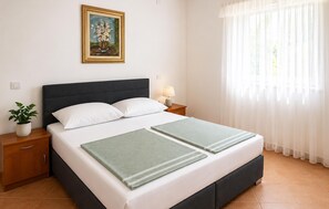 Ein ordentlich bezogenes Bett mit zwei Kissen, ein Nachttisch mit Lampe, ein gerahmtes Bild an der Wand und ein Fenster mit durchsichtigen Vorhängen.