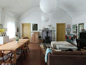 A room with a wooden dining table, chairs, a sofa, a fireplace, and a large window. - Old parish hall in Kvæfjord (Borkenes)