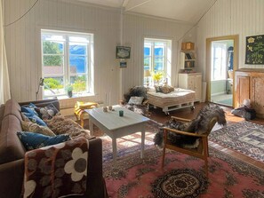 A cozy living room with a sofa, armchair, and a small table. There are two windows with curtains, a fireplace, and a bookshelf.