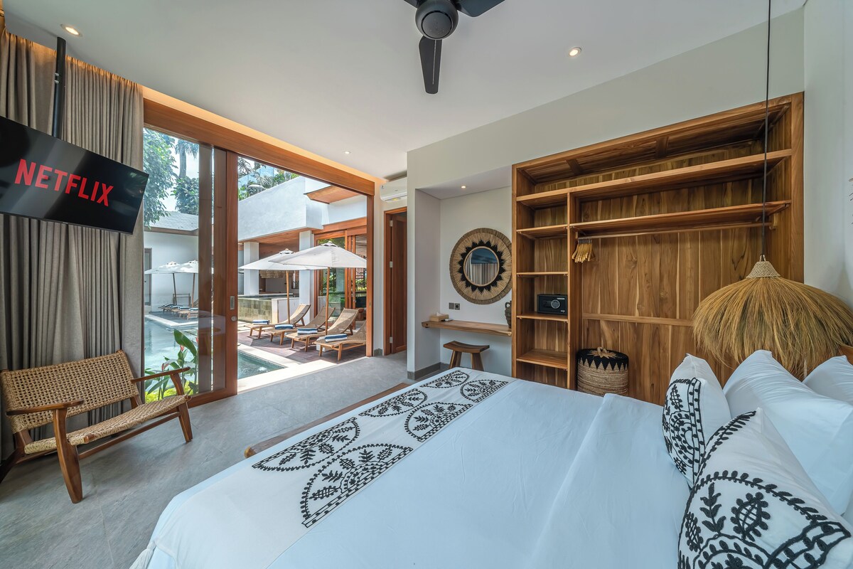 Ein modernes Hotelzimmer mit einem großen Bett, einem Fernseher, einer Holzbücherregal und Blick auf den Außenpoolbereich.