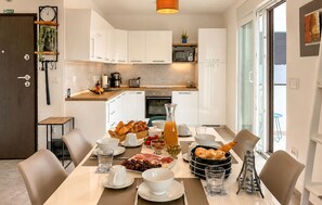 Eine moderne Küche mit weißen Schränken, einer Mikrowelle und einer Kaffeemaschine. Ein Esstisch ist gedeckt mit Tellern, Gläsern und verschiedenen Speisen.