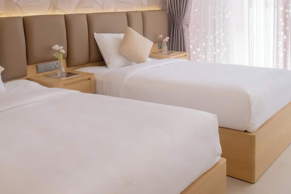 A hotel room with two beds, a large window with sheer curtains, a wall with a leaf pattern, and a nightstand with a vase and a book.