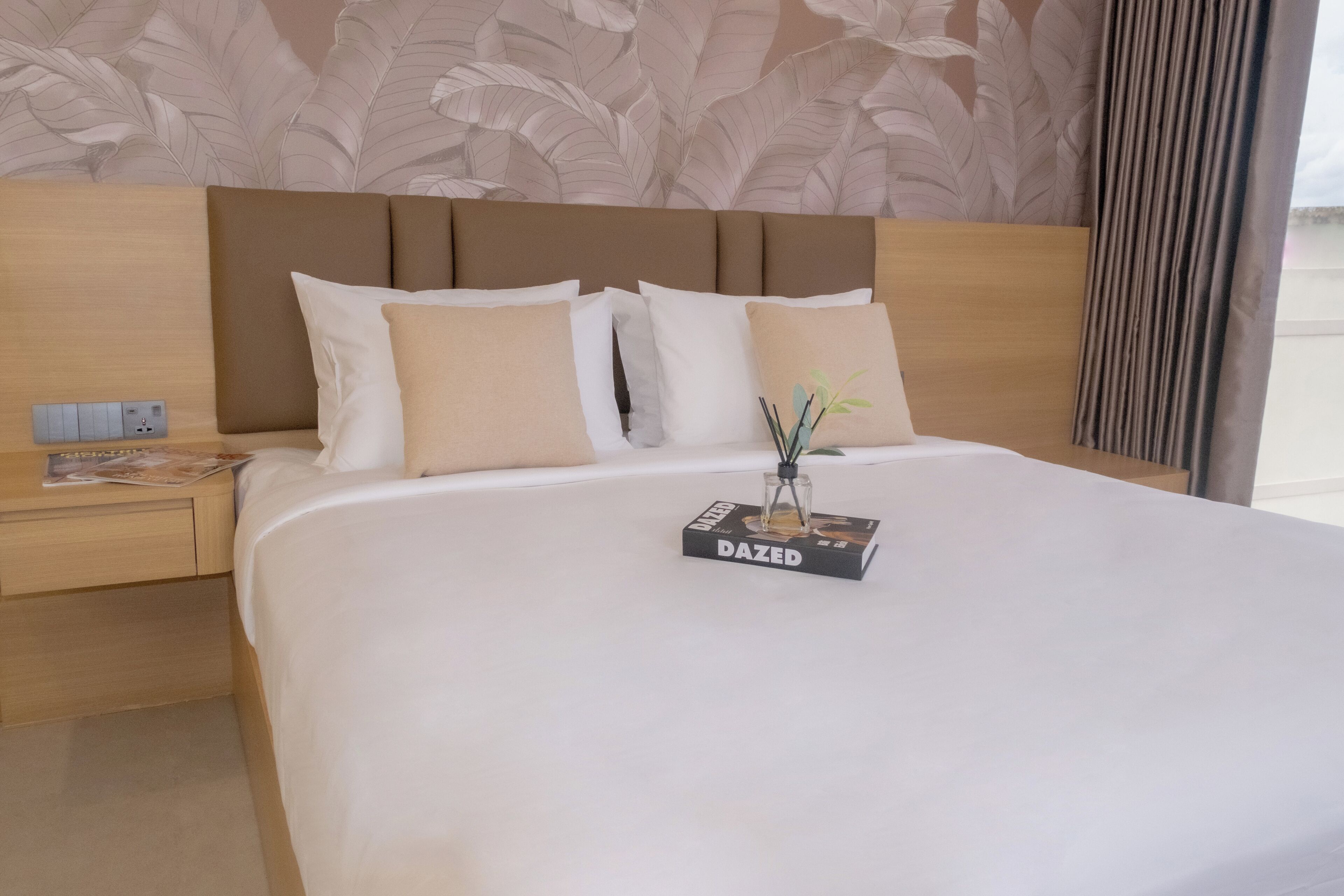 A neatly made bed with a white comforter and several pillows, a bedside table with a magazine, and a wall with a leaf pattern.