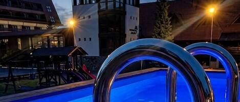 Una piscina con dos copas de champán y un farol, rodeada por una terraza de madera y un edificio con una torre.