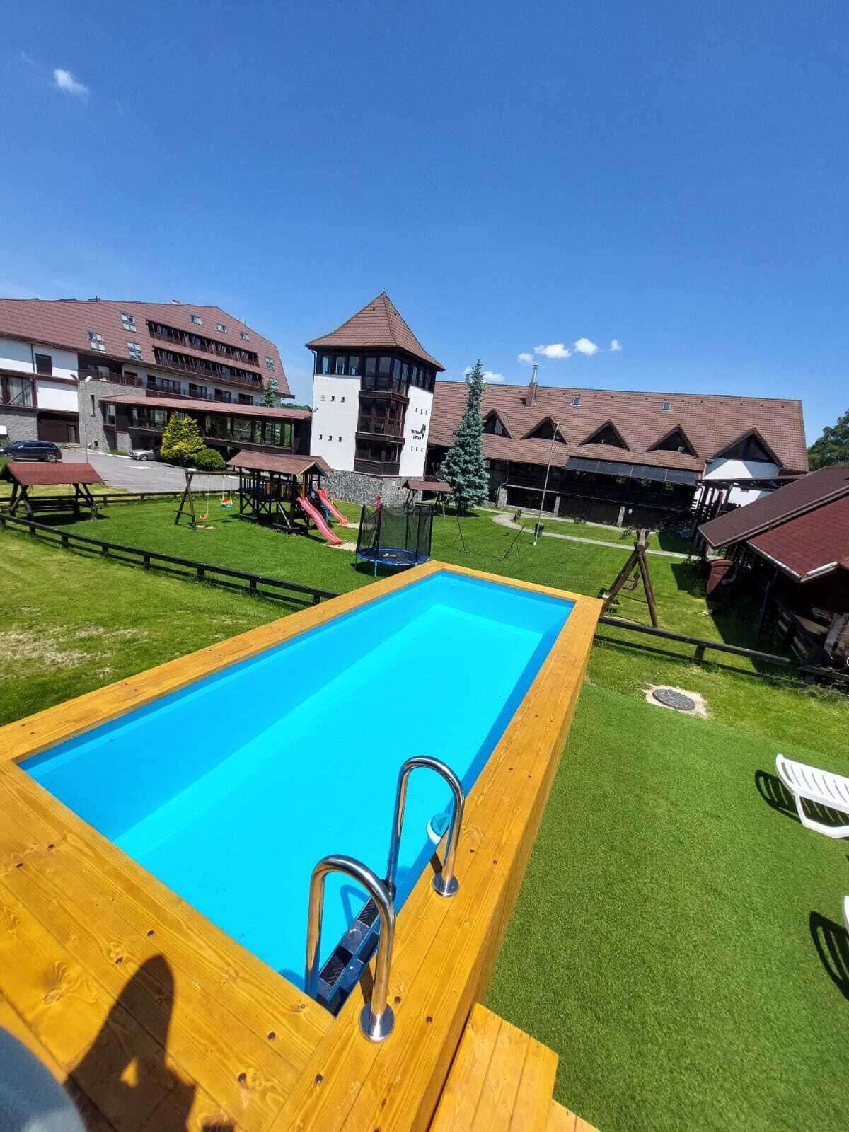 Une piscine avec une terrasse en bois, un espace de jeux pour enfants et un bâtiment doté de nombreuses fenêtres et d’une tour.