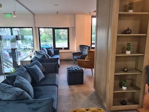 Un interior moderno con un sofá, sillones, una estantería de madera y vistas a la calle a través de grandes ventanales.