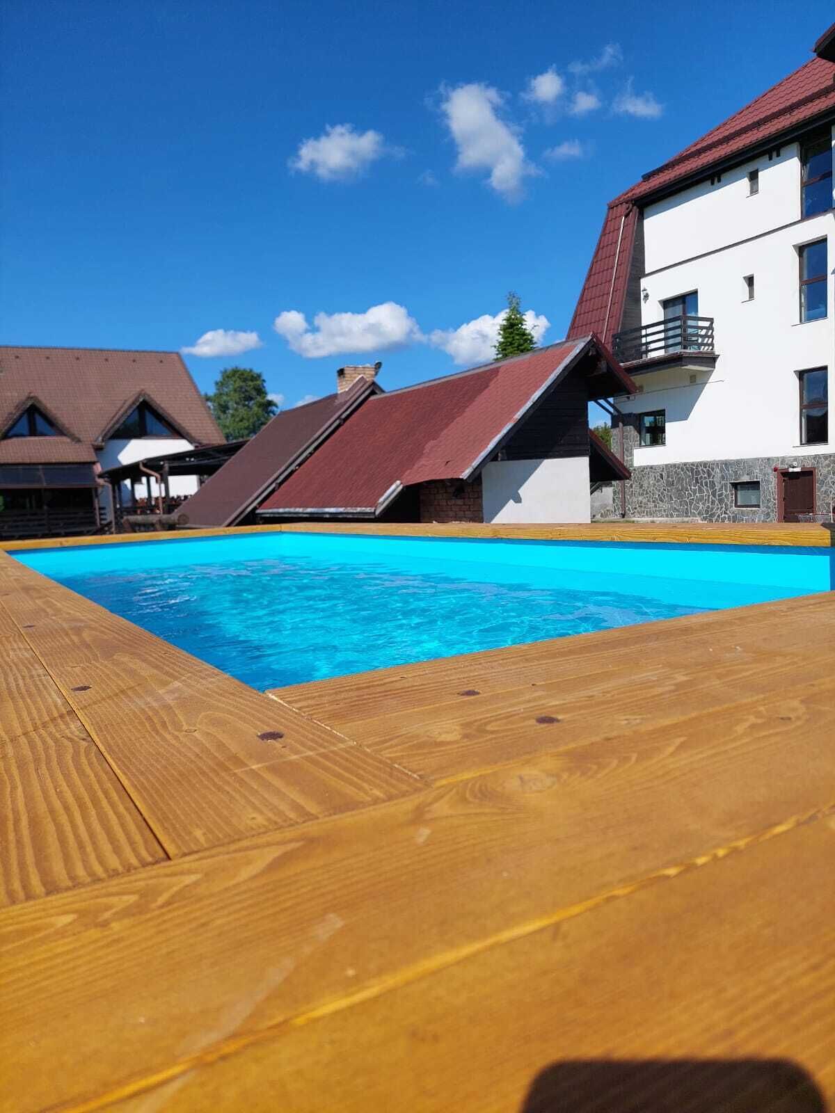 Une terrasse en bois mène à une piscine, avec un ciel bleu dégagé et des bâtiments en arrière-plan.