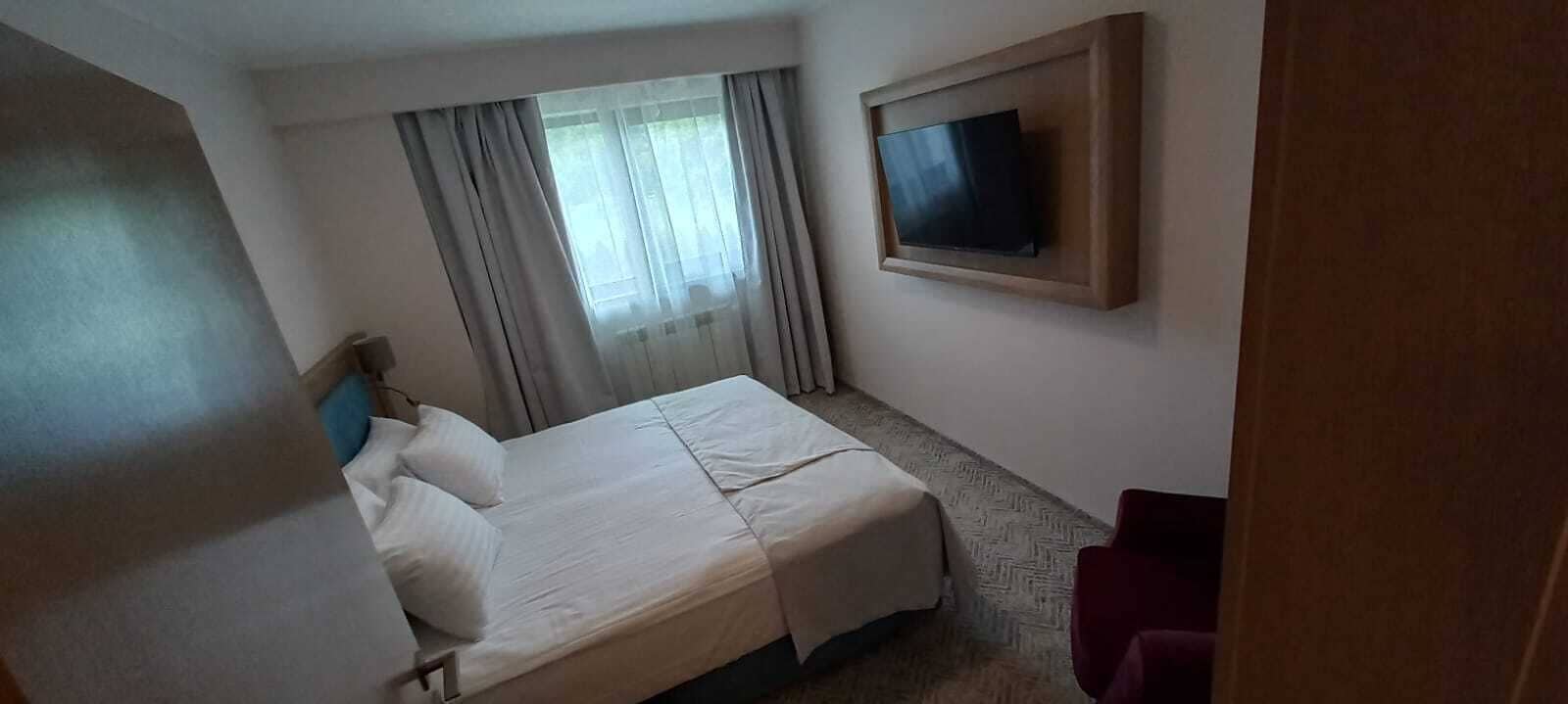 A hotel room with a bed, a TV mounted on the wall, a window with curtains, and a red chair.