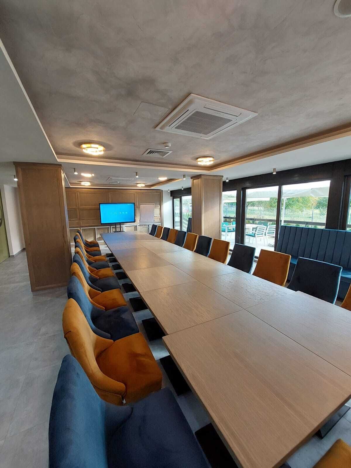 Ein Konferenzraum mit einem langen Tisch, mehreren Stühlen, einem Fernsehbildschirm und großen Fenstern.