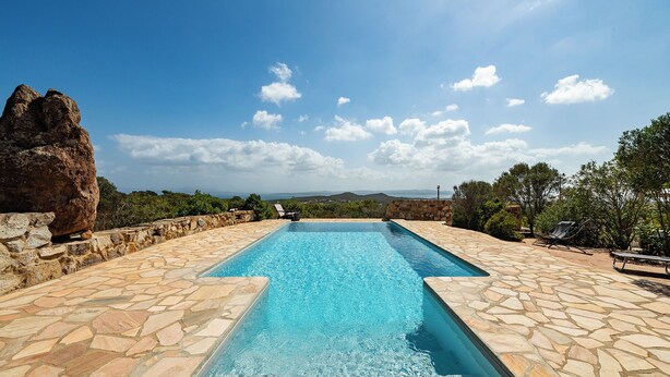 A rectangular pool with stone flooring, surrounded by a stone wall and vegetation, with a clear sky and distant landscape.