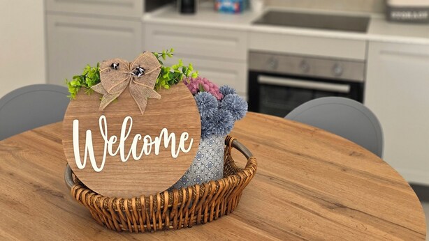 Una mesa redonda de madera con una cesta de mimbre que contiene un cartel de bienvenida, un florero con flores y una tetera sobre una encimera de cocina moderna.