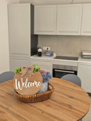 Une table ronde en bois sur laquelle repose un panier en osier contenant un panneau d’accueil, un vase avec des fleurs et une bouilloire, le tout placé sur un plan de travail moderne dans la cuisine.