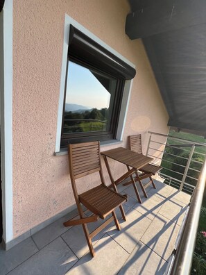 A balcony with two wooden chairs and a table, a window with a view of greenery, and a textured wall. - Spacious Alpine Retreat | 3BR, 2BA & Wood-Fired Sauna (Kobarid)