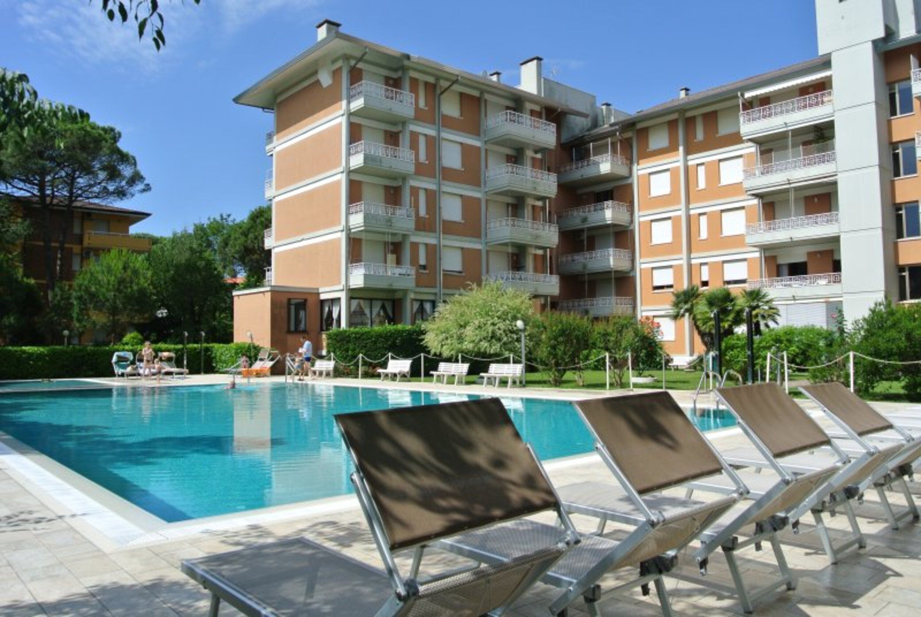 A swimming pool with lounge chairs, a multi-story building with balconies, and greenery in the background.