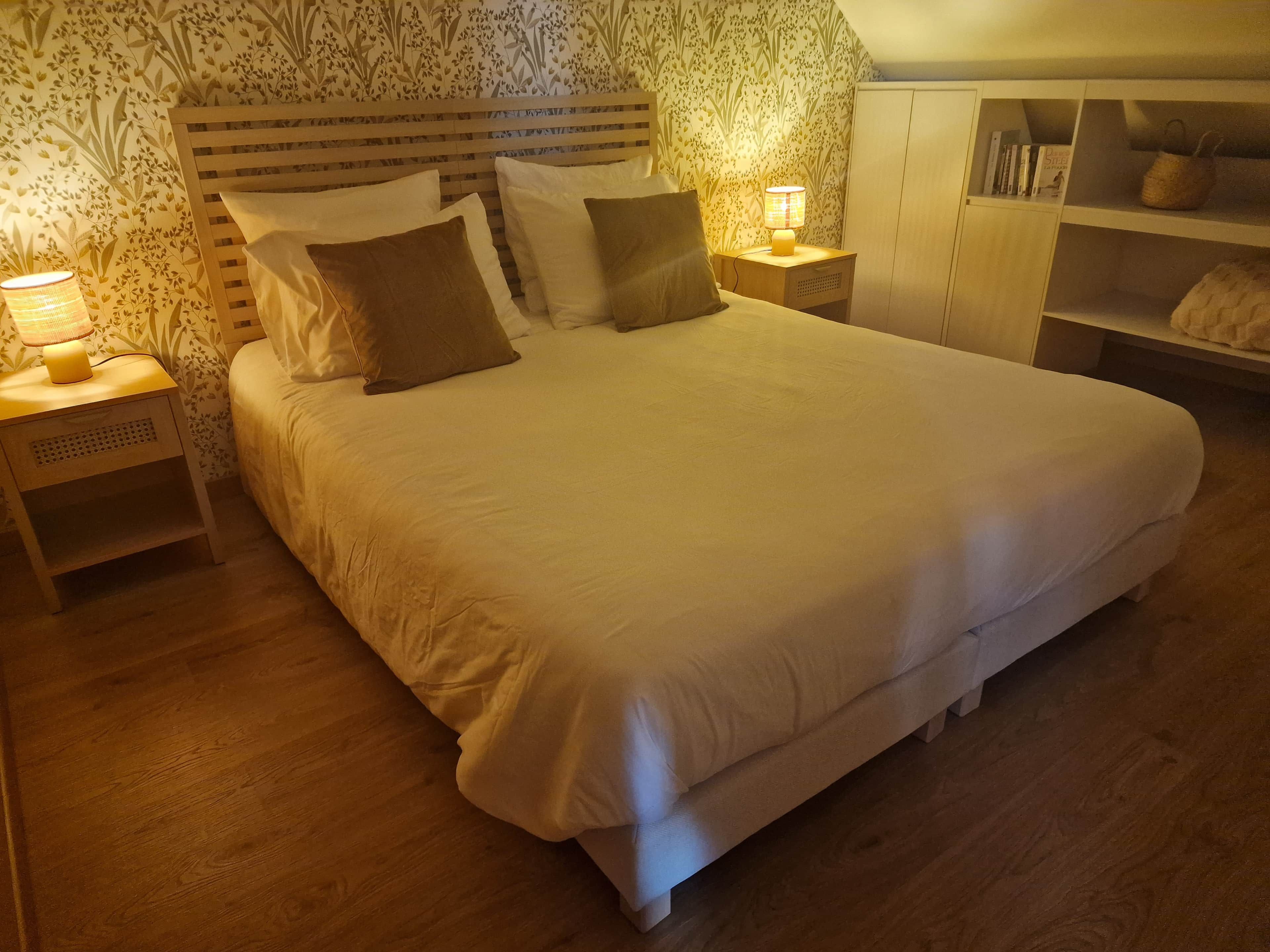 Ein ordentlich bezogenes Bett mit weißer Bettwäsche und mehreren Kissen, seitlich von zwei Nachttischen mit Lampen, in einem Zimmer mit Holzboden und strukturierten Tapeten.