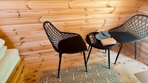 Interior de una cabaña de madera con dos sillas de malla negra, una mesita y un tapete estampado.