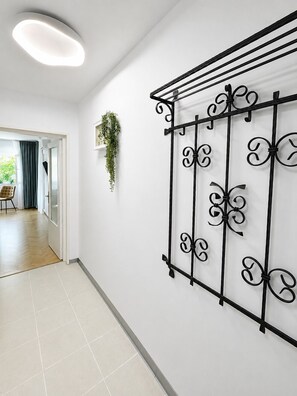 Ein Flur mit weißer Wand, einer schwarzen Metall-Dekoration, einer hängenden Pflanze und einer Tür zu einem anderen Raum.