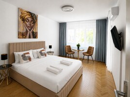 Habitación de hotel con cama, un cuadro, televisor, escritorio, dos sillas y ventana con cortinas.