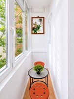 Un pasillo estrecho con una ventana, un cuadro enmarcado, una mesita con una planta y dos sillas color naranja.