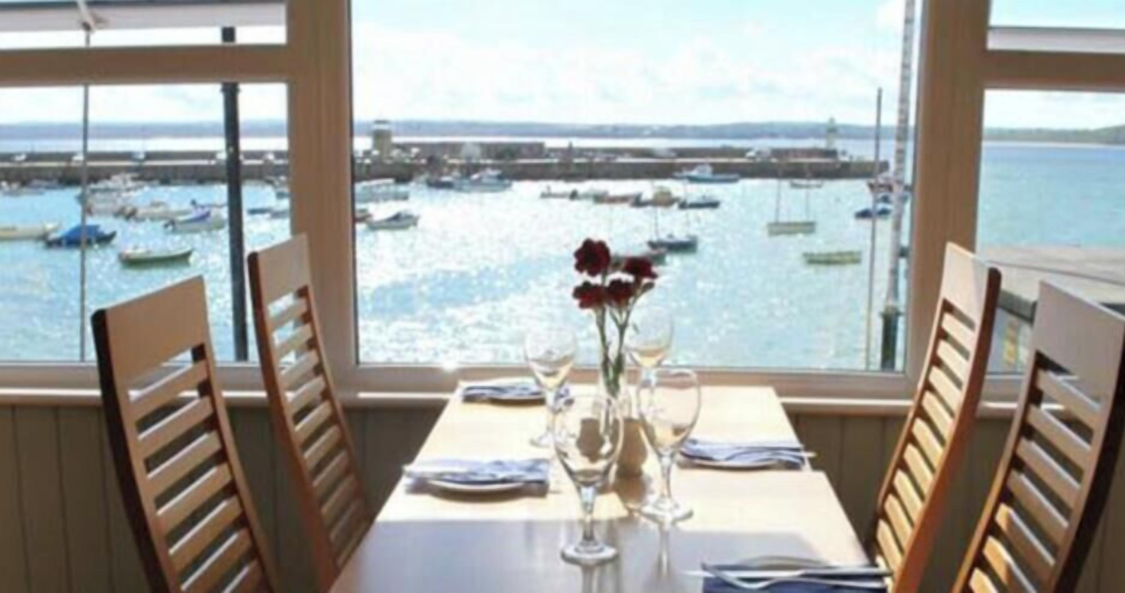 Una mesa de comedor con sillas, un florero con flores y vistas a barcos en un puerto a través de ventanas amplias.