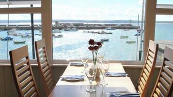 Una mesa de comedor con sillas, un florero con flores y vistas a barcos en un puerto a través de ventanas amplias.