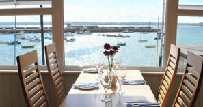 A dining table with chairs, a vase with flowers, and a view of boats in a harbor through large windows.