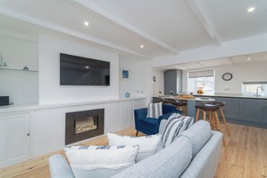 A modern living room with a fireplace, a TV mounted on the wall, and a kitchen area in the background.