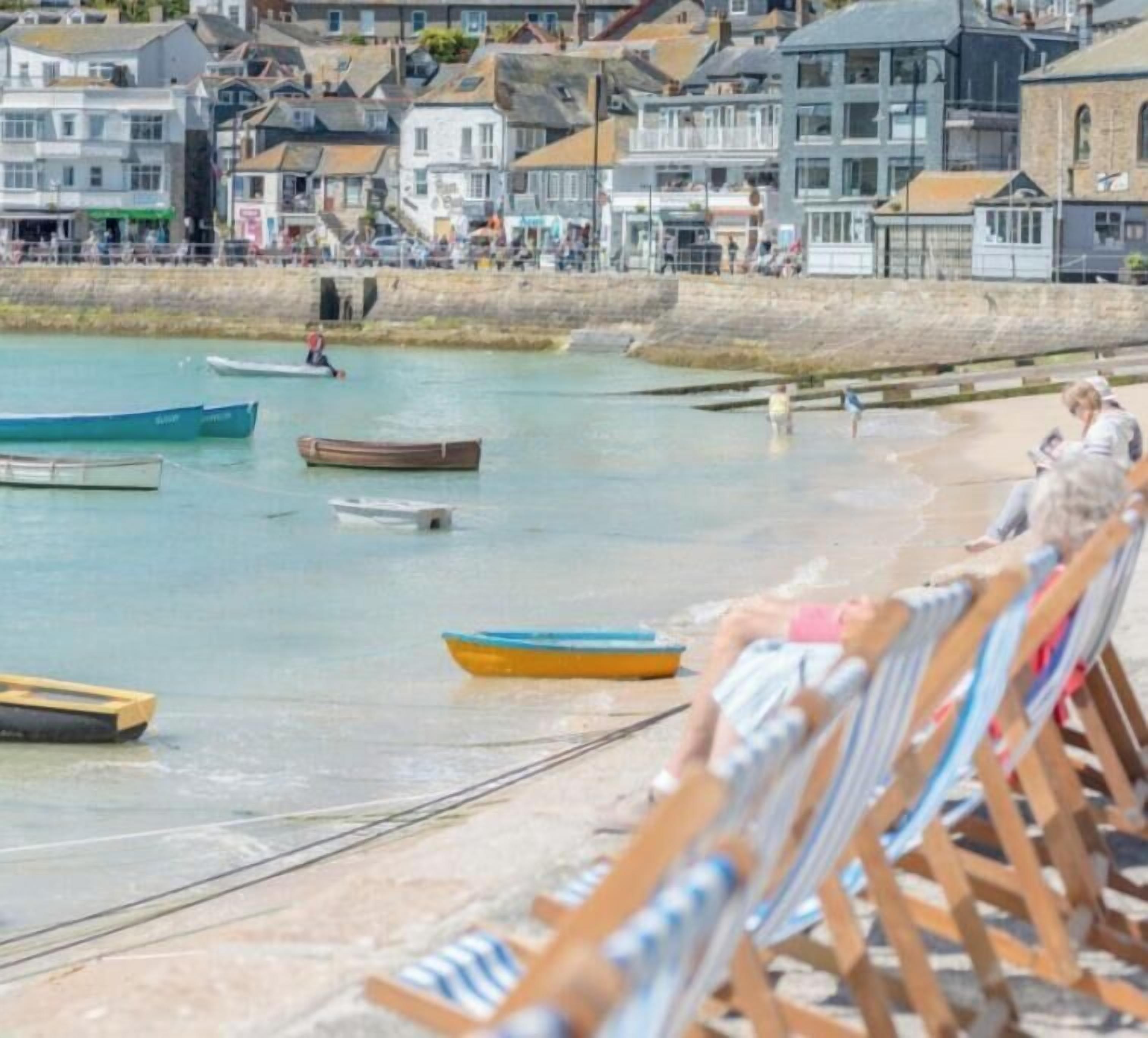 Paisaje costero con barcos en el agua, una playa con tumbonas de madera y edificios al fondo.