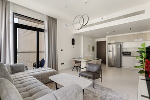 Ein modernes Wohnzimmer mit einer Eckcouch, einem Esstisch mit Stühlen, einer Küchenzeile mit Kühlschrank und Mikrowelle sowie einem großen Fenster mit Vorhängen.