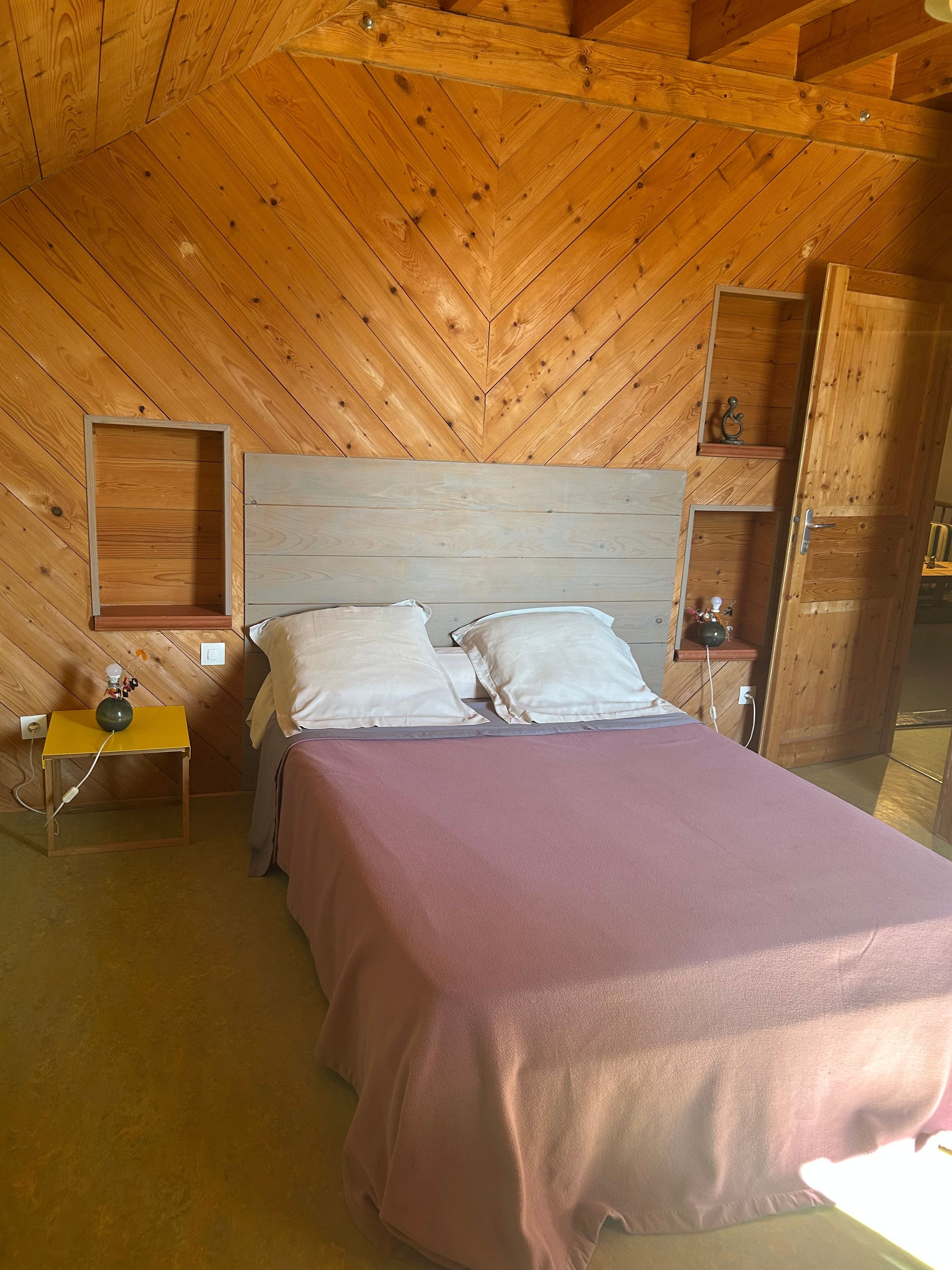 Una camera da letto con soffitto in legno, un letto con copripiumino rosa, due cuscini bianchi, un comodino con una lampada e uno scaffale con una bottiglia.
