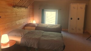 Une pièce aux murs en bois, avec un plafond en pente, deux lits, une fenêtre, une armoire et une lampe.
