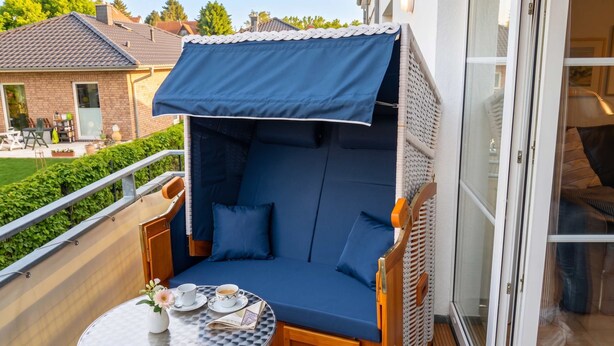 A balcony with a blue cushioned seating area, a small table with cups and a flower vase, and a view of a house and greenery. - FeWo Frische Brise - Lauterbach (Putbus)
